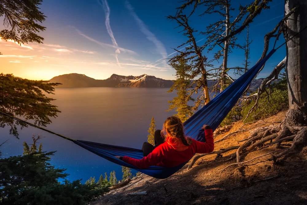Woman reclining in a hammock looking over Crater Lake with the sun setting behind snow-covered mountains