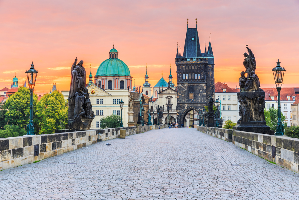 A view from the perspective of someone walking along the path on top of the Charles Bridge during dusk with the sun setting behind the Old Town Tower during the overall best time to go to Prague
