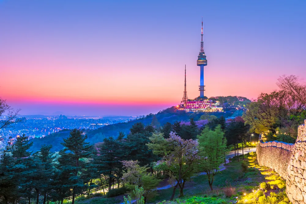 the Seoul Tower pictured in Spring, the best time to visit South Korea, with green trees and the city lit up in multi-colored lights at night
