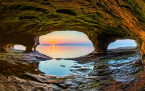 Neat view of a sea cave pictured during the best time to visit the Upper Peninsula featuring a sunset over the water, as viewed from inside the cave