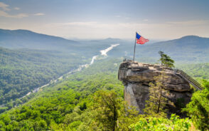 Chimney Rock, one of the best places to visit in North Carolina, pictured towering over the forest below with an American flag flying high above the staircase and rock face on a hazy day