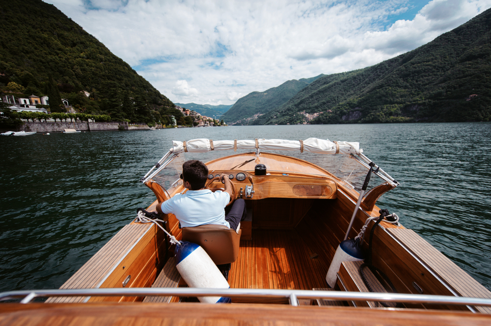 Antique wooden boat driving along the water between big hills on either side during the best time to visit Lake Como