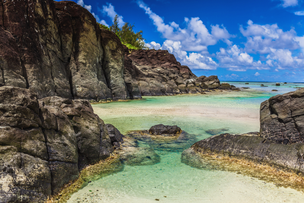 Gorgeous view of black rocks surrounding a beach during the best time to visit the Cook Islands with blue skies overhead