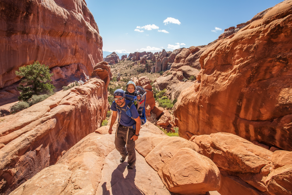 A father carrying his son in a backpack up a rock face during the least busy time to visit Moab