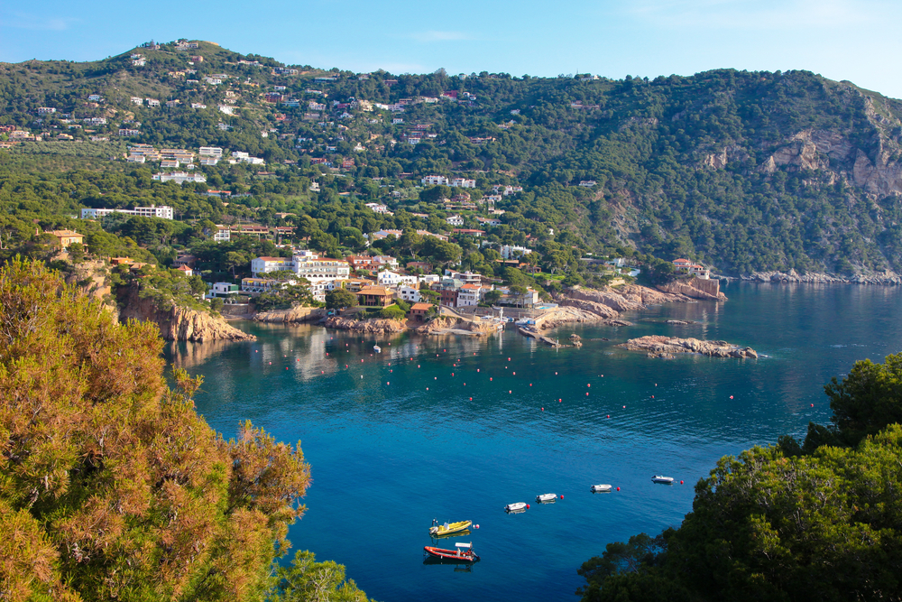 Idyllic town of Begur pictured from the top of a hillside overlooking the beach and boats and resorts below, as seen from between a couple trees