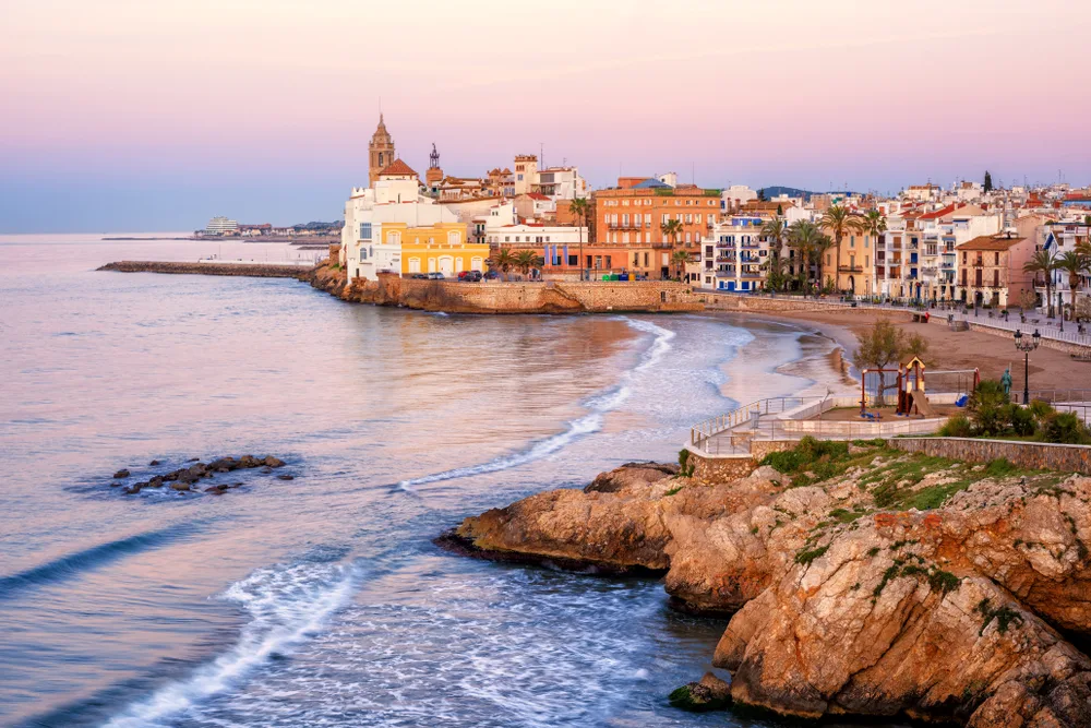Seaside resort of Sitges pictured at dusk with waves lapping the rocky coastline for a piece on the best day trips from Barcelona