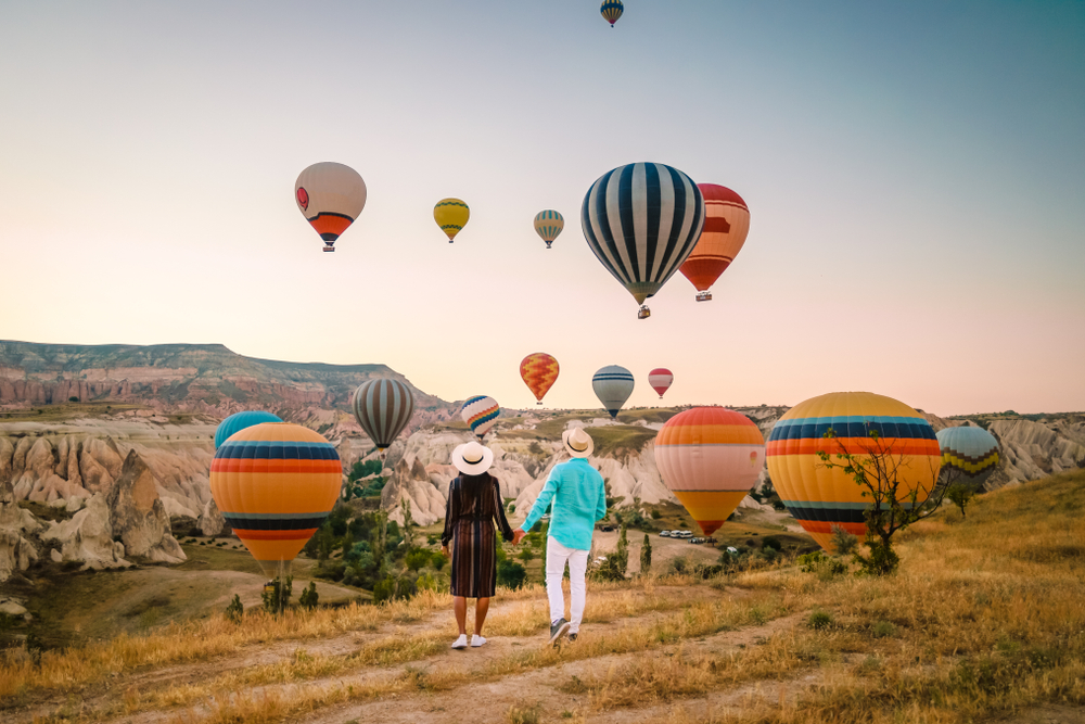 Happy couple holding hands and watching the sun rise while balloons rise up from the valley during the best time to visit Cappadocia