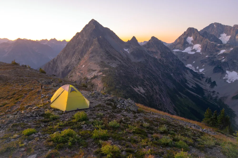 Lone camper using a yellow tent on the side of a mountain, pictured at dusk with the sun setting over the mountains