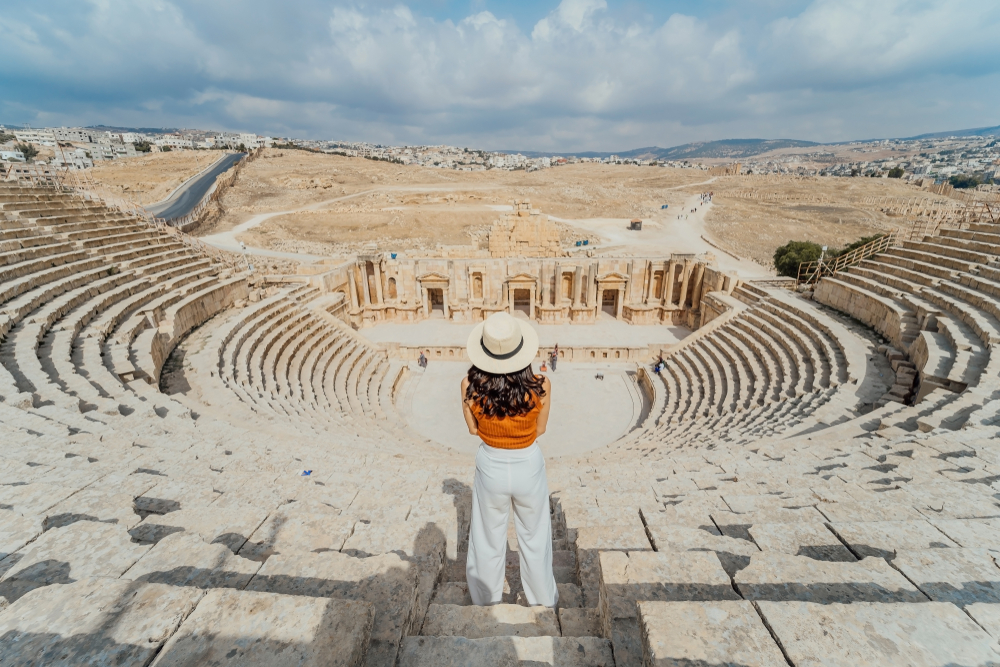 Woman in white p ants and a beige hat pictured standing on the steps of an amphitheater overlooking some ruins in Jordan