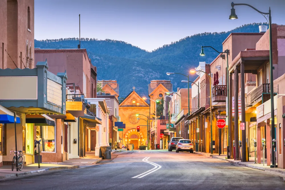 Neat dusk view of downtown Santa Fe, one of the best places to visit in the US, pictured with lights on the walkways and the mountains in the backgrounds