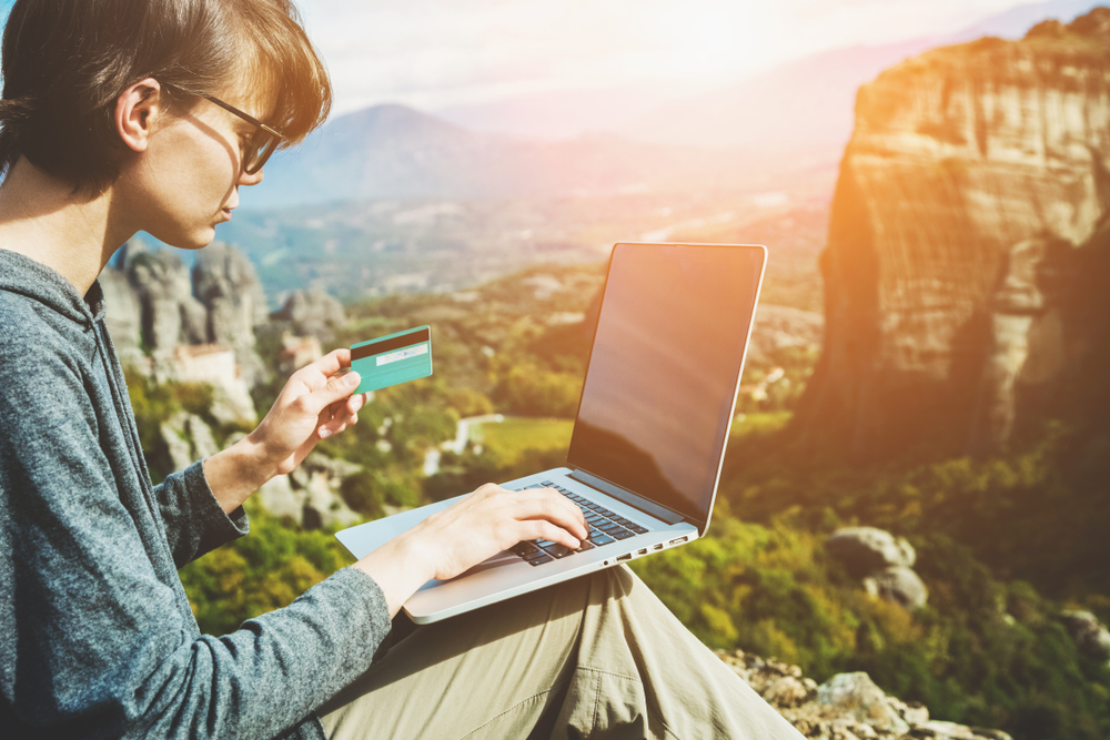 Woman sits outdoors overlooking mountains with laptop and credit card on her knee for a piece on the best time and day to book flights