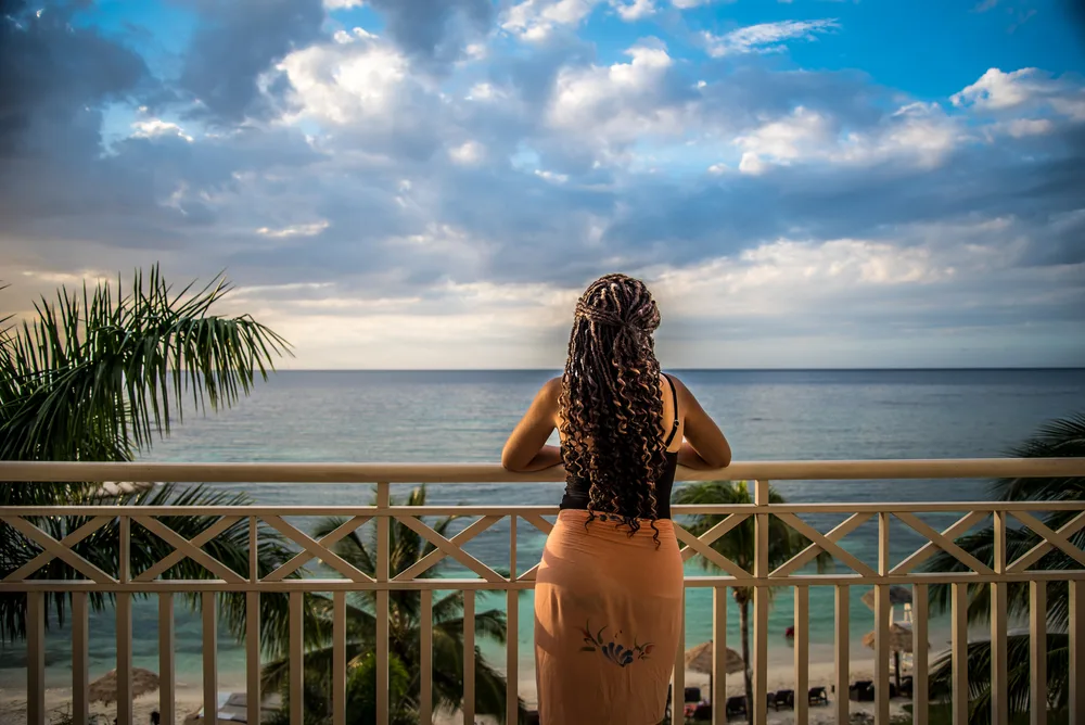 Woman in a peach-colored dress pictured from the back looking out over the ocean with clouds in the sky