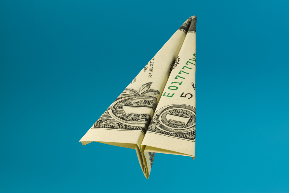 Folded dollar bill in a paper airplane shape against a blue background for a piece on the cheapest days to fly