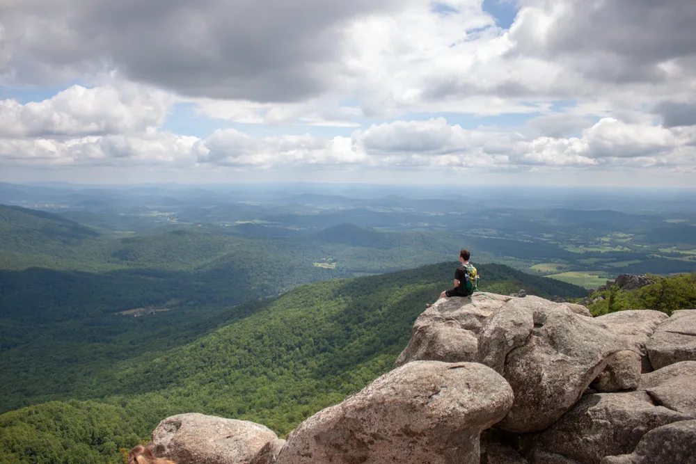 Man sitting on a rock face that's protruding out from a tall cliffside overlooking a green forest below during the best time to visit Shenandoah National Park
