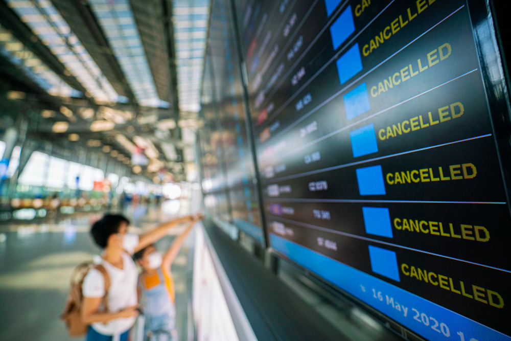 Asian woman stands in front of flight status monitor showing cancelled flights for a piece on what is travel insurance