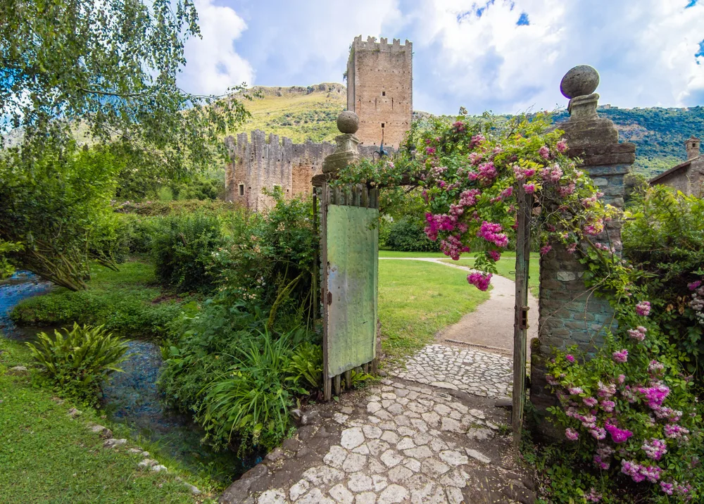 Ninfa Gardens pictured through a stone and wooden gate with a castle-like structure in the background