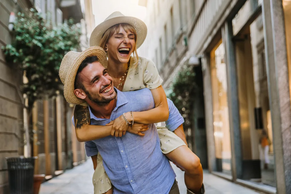Cute couple in hats laughing while they travel internationally, pictured in the street for a piece on the best travel quotes for couples
