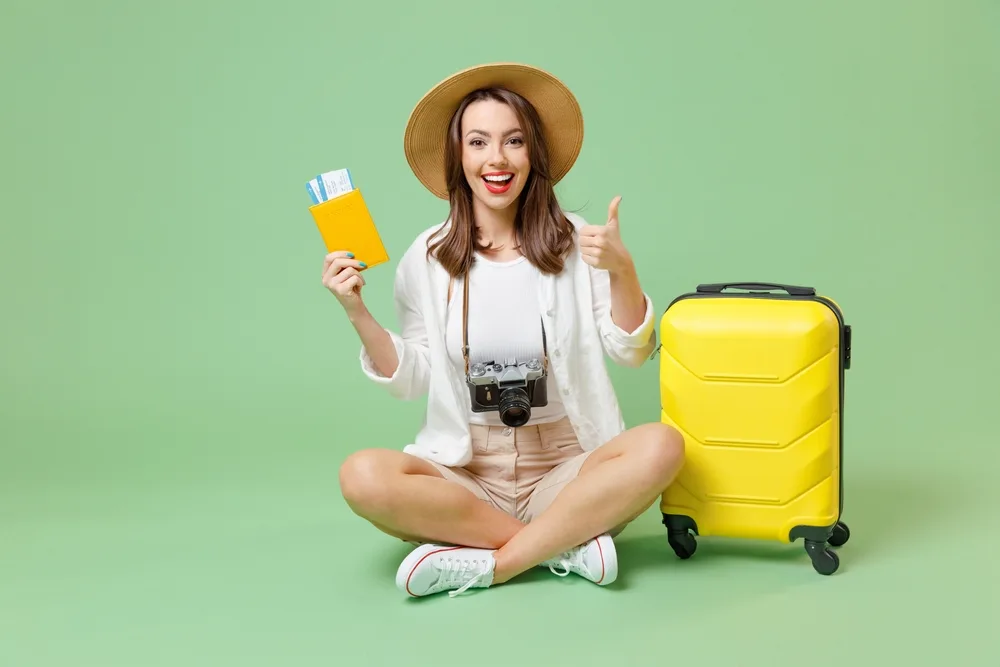Woman sitting cross-legged on the ground in a green room with a yellow suitcase giving the camera a thumbs-up