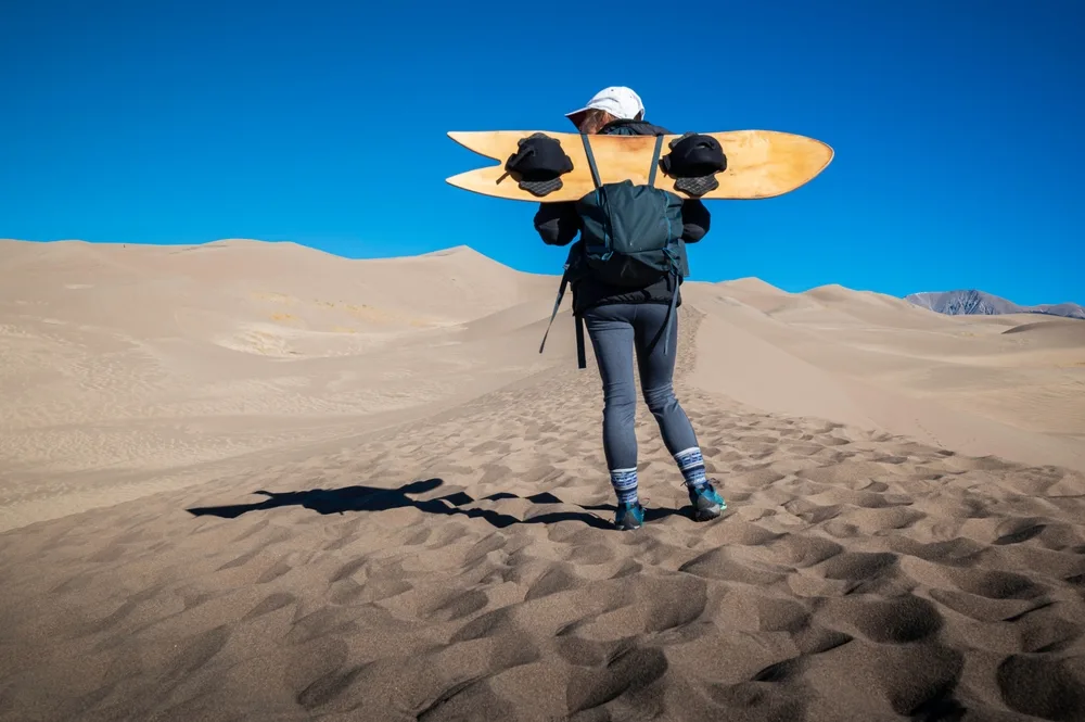 Woman holding a sandboard on her shoulders behind her and looking left while wearing yoga pants and a backpack