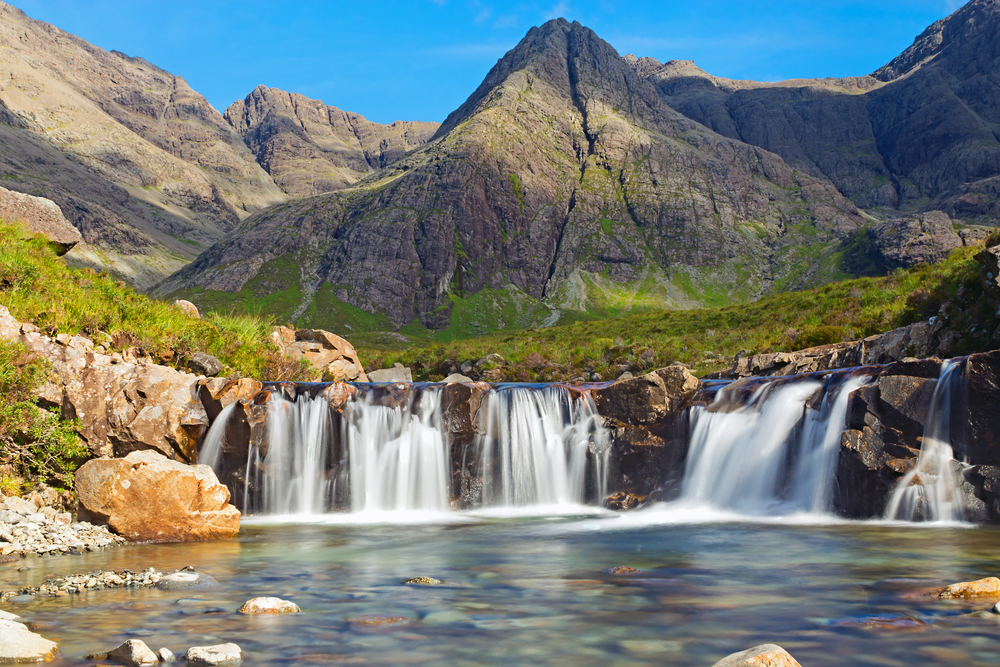 The Skye Fairy Pools, a top pick for must-stt places to visit in Scotland, pictured with rolling green hills in the background and the falls in the foreground