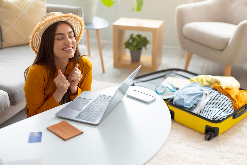 Young woman with straw hat expresses joy in front of her laptop after booking a flight with a yellow packed suitcase nearby