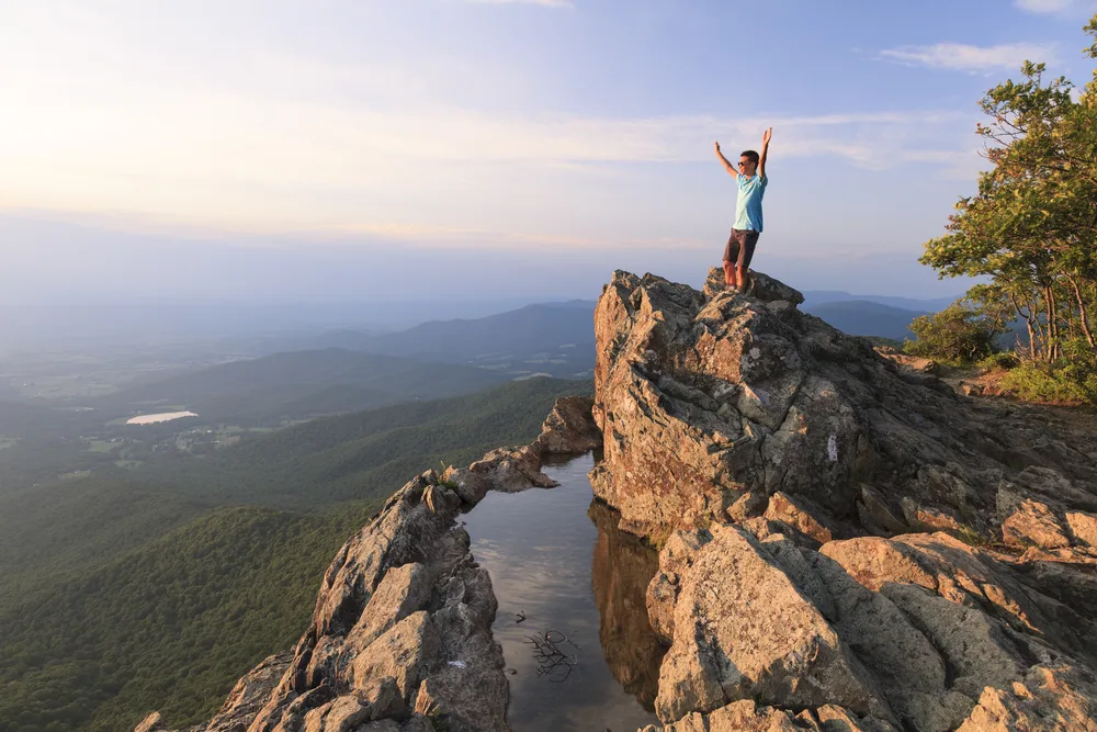 Guy standing on a rock face and holding his arms up with a natural pool below him that overlooks a dense forest in Shenandoah National Park