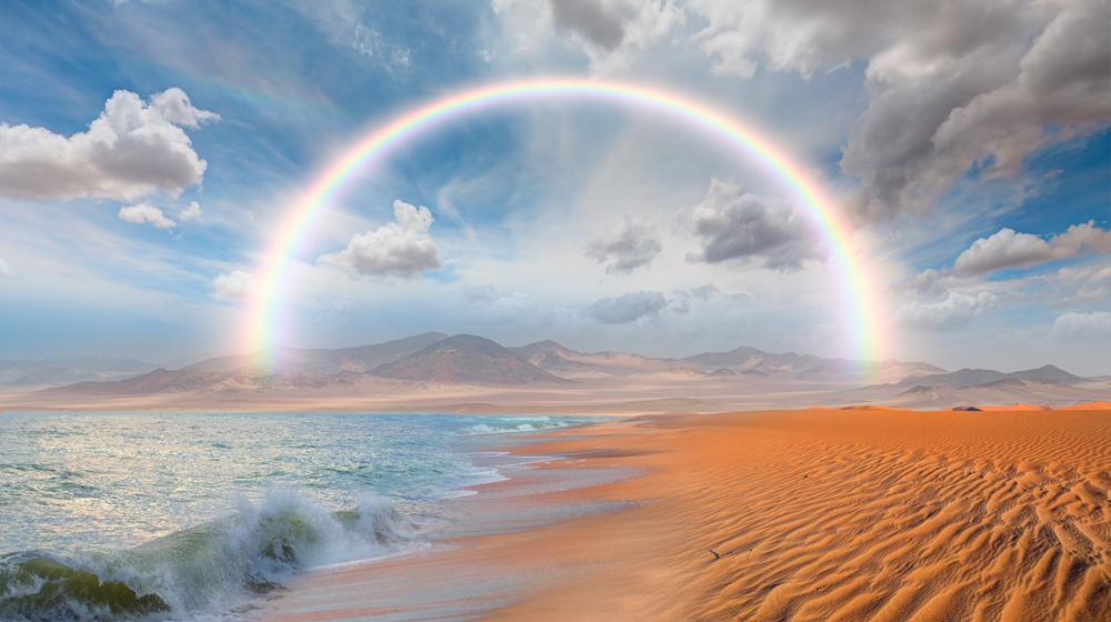 Desert in Namibia pictured with a rainy sky and a rainbow over where the ocean and desert meet