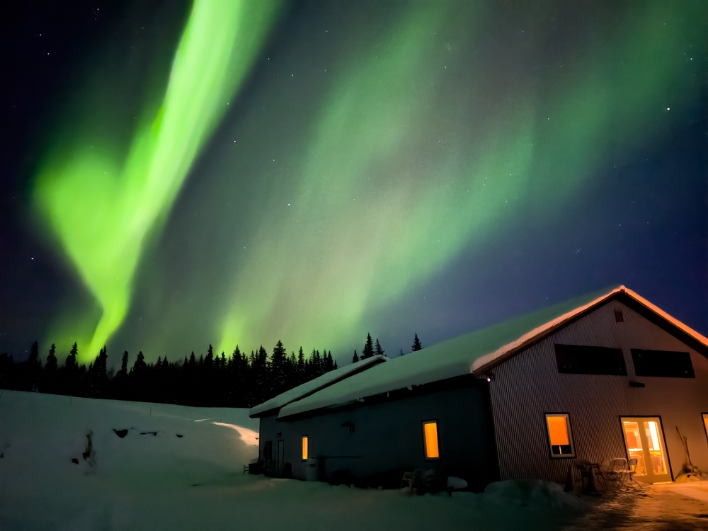 Green Northern Lights seen over a snow-covered house on a winter day during the overall cheapest time to visit Fairbanks