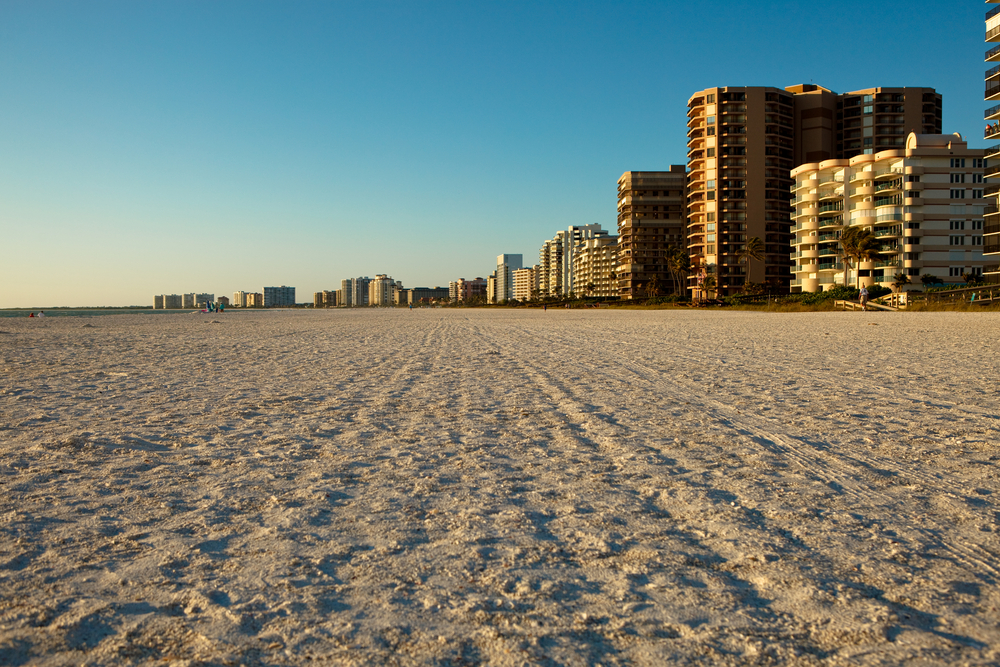 For a guide to the best time to visit Marco Island, a gorgeous view of the beige sand and small condo buildings overlooking it below a blue sky