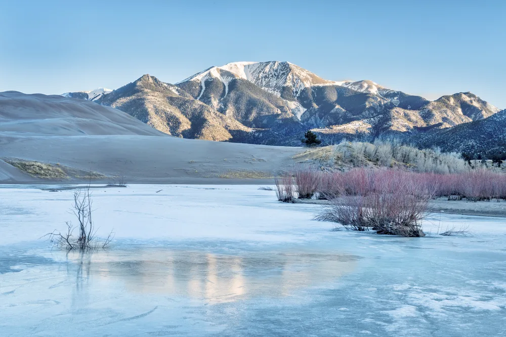 Frozen lake pictured in the foreground with snow-capped dunes in the winter, the overall cheapest time to visit the Great Sand Dunes National Park