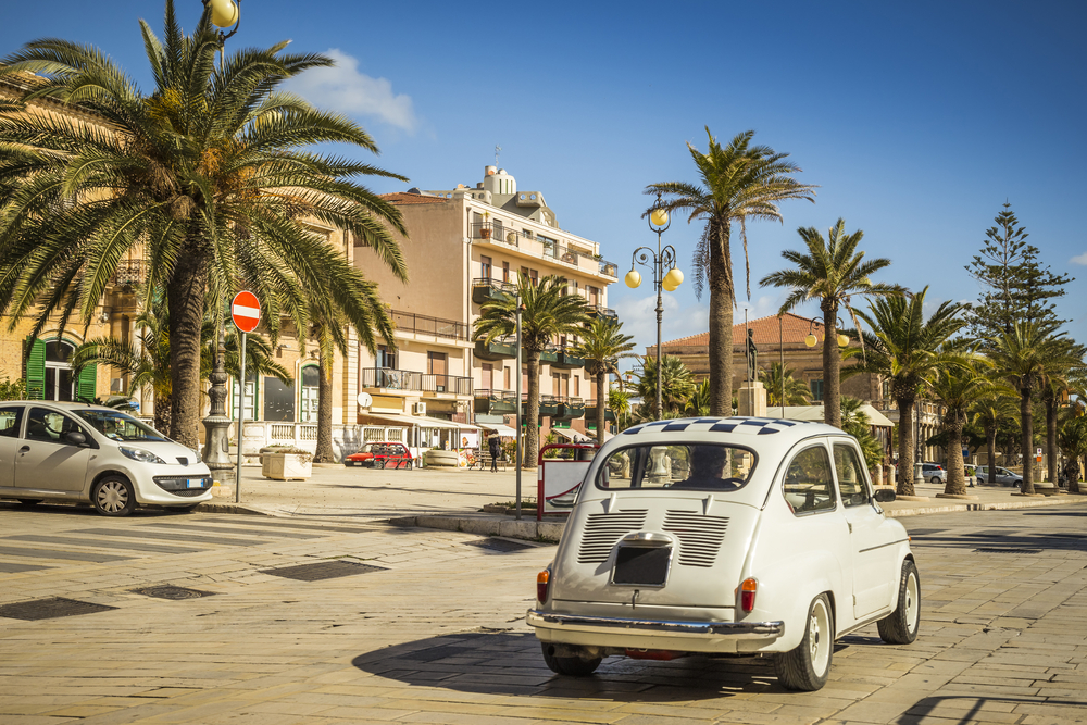 Fiat on a stone street pictured during the least busy time to visit Sicily with palm trees in the middle of stone homes