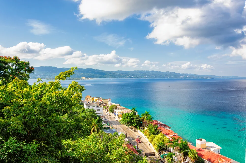 Gorgeous view of the ocean and quaint homes and shops along the bay with vegetation on the left of the road on a clear day during the best time to visit Montego Bay