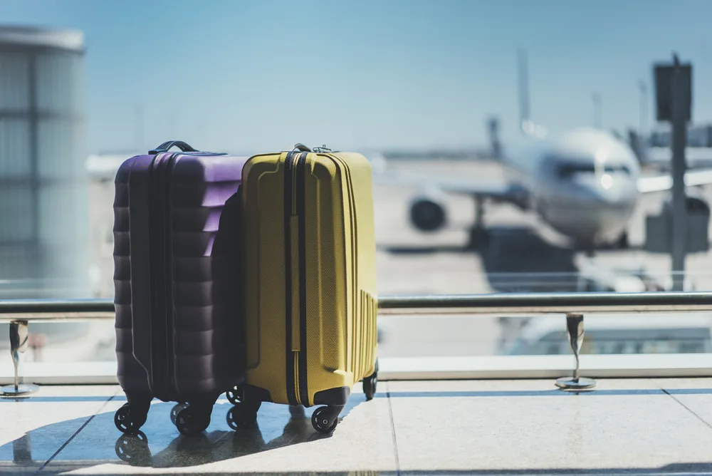 Yellow and purple hardbody suitcases sit in front of a window at an airport for a piece asking how much does travel insurance cost?