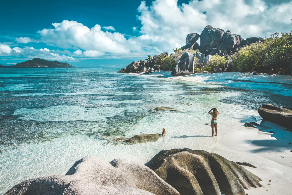 Fit woman in a white bikini taking a photo of a rock formation while standing in ankle-deep water during the best time to visit Seychelles