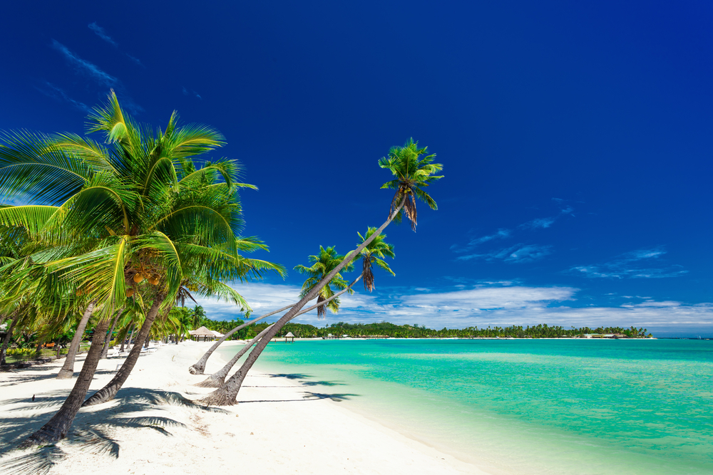 Palm trees lie over a gorgeous white sand beach and teal water on Plantation Island in Fiji, a perfectly safe place to visit