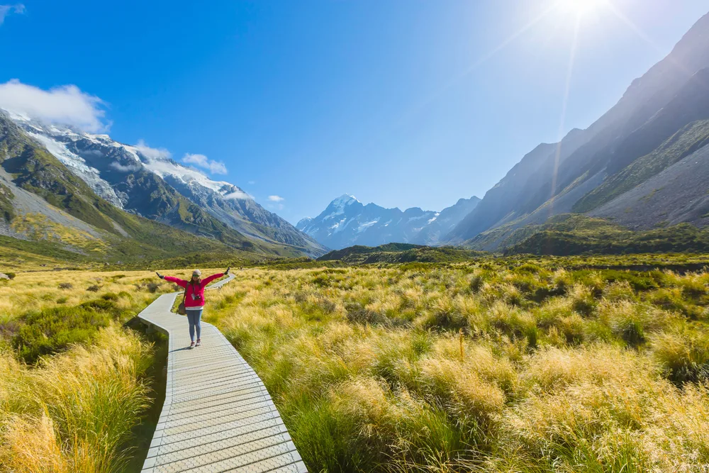 Asian woman holding her arms above her head while standing on a boardwalk in Mount Cook National Park in New Zealand in a photo that's prime for the best travel quotes