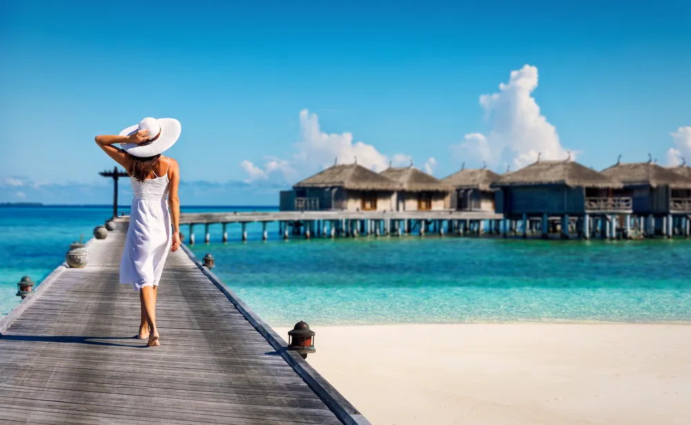 Woman in a white dress and a white hat holding her hat and walking on a wooden walkway leading to some overwater bungalows in the Maldives