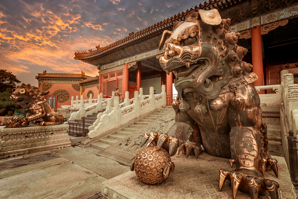 Lion statue occupies the foreground of the Forbidden City, one of the best places to visit in the World, with an orange sky as seen at dusk