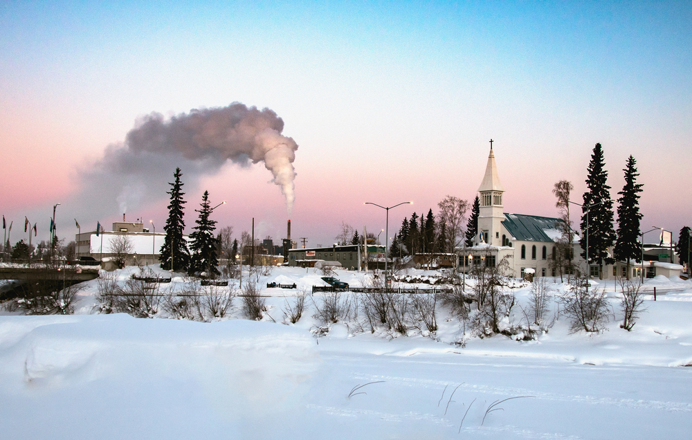 Old white church pictured from across a frozen plain during the winter, the overall worst time to visit Fairbanks, with smoke rising up into the sky from a stack