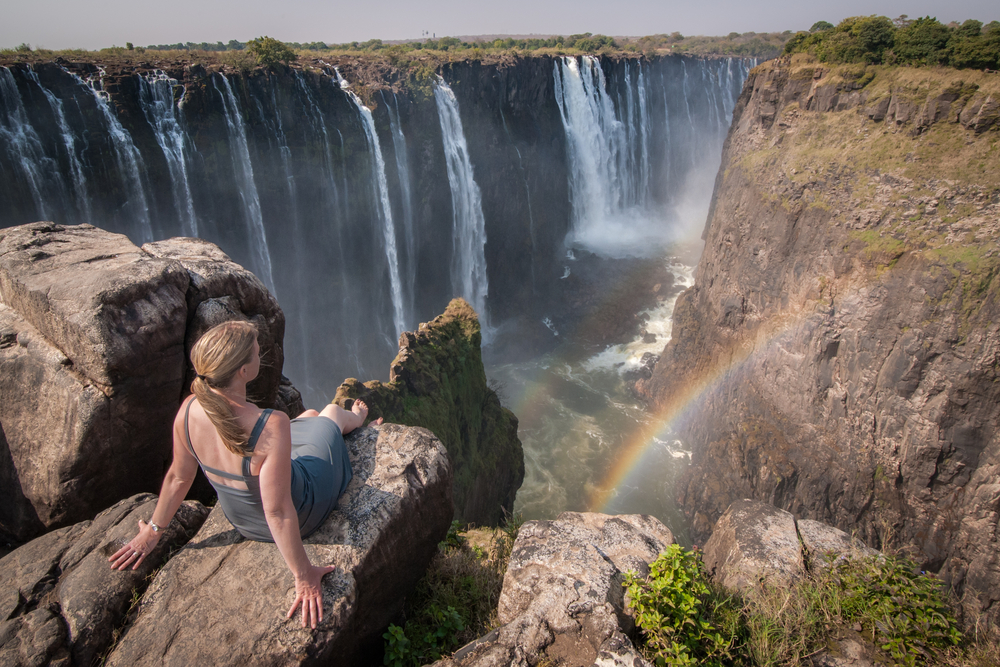 Woman in a grey dress sitting on a dry rock overlooking the Falls with a rainbow below during the best time to visit Victoria Falls