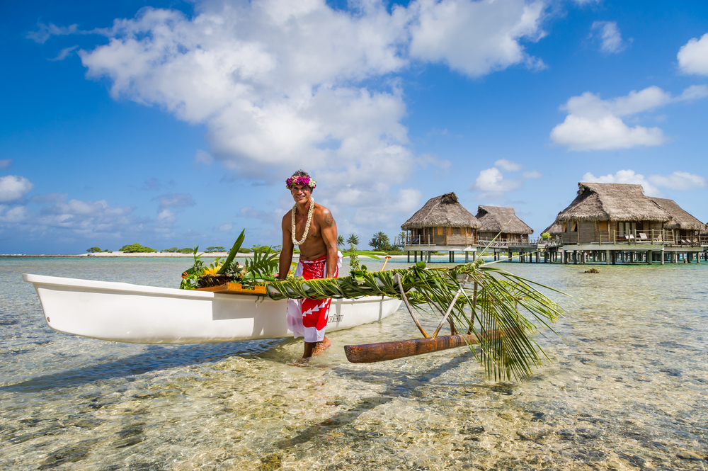Local Tahitian man wearing flowers on his head pictured during the best time to visit French Polynesia with huts in the background and vegetation on his boat