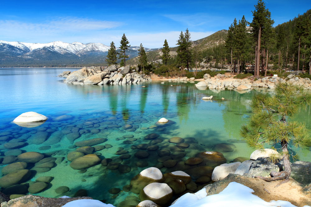 Rocks on the shore of Lake Tahoe, pictured with white rocks protruding up from the crystal-clear water and snow-capped mountains in the background