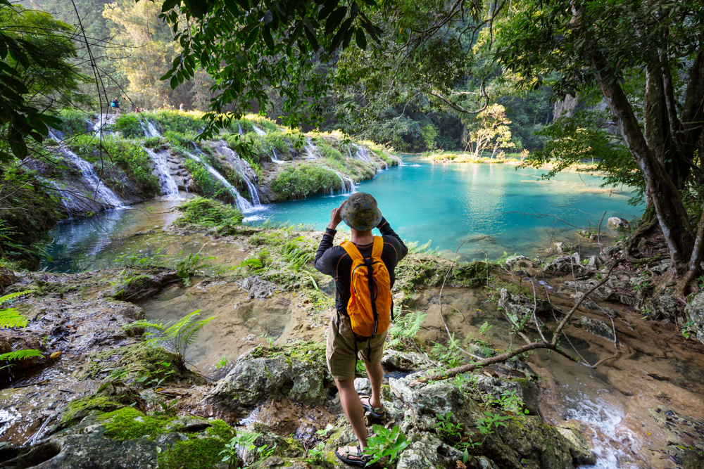 Asian man photographing the natural pools at Semuc Champey, one of the best places to visit in Guatemala, as seen from the trees with water flowing over the tiered sections in little falls