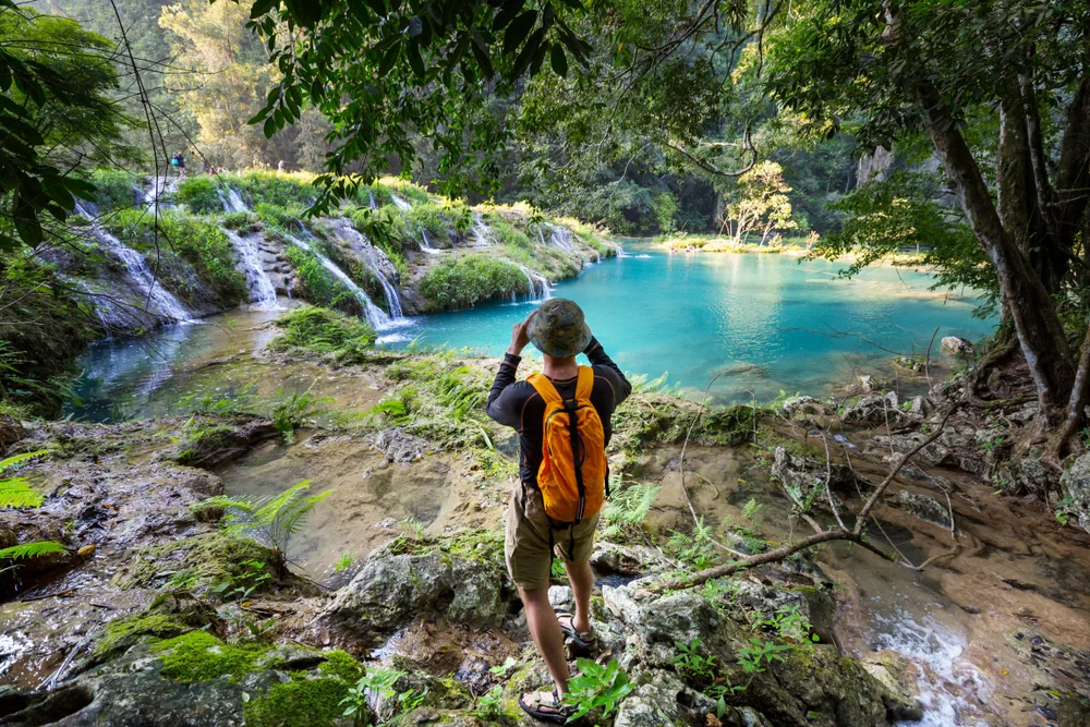 Asian man photographing the natural pools at Semuc Champey, one of the best places to visit in Guatemala, as seen from the trees with water flowing over the tiered sections in little falls