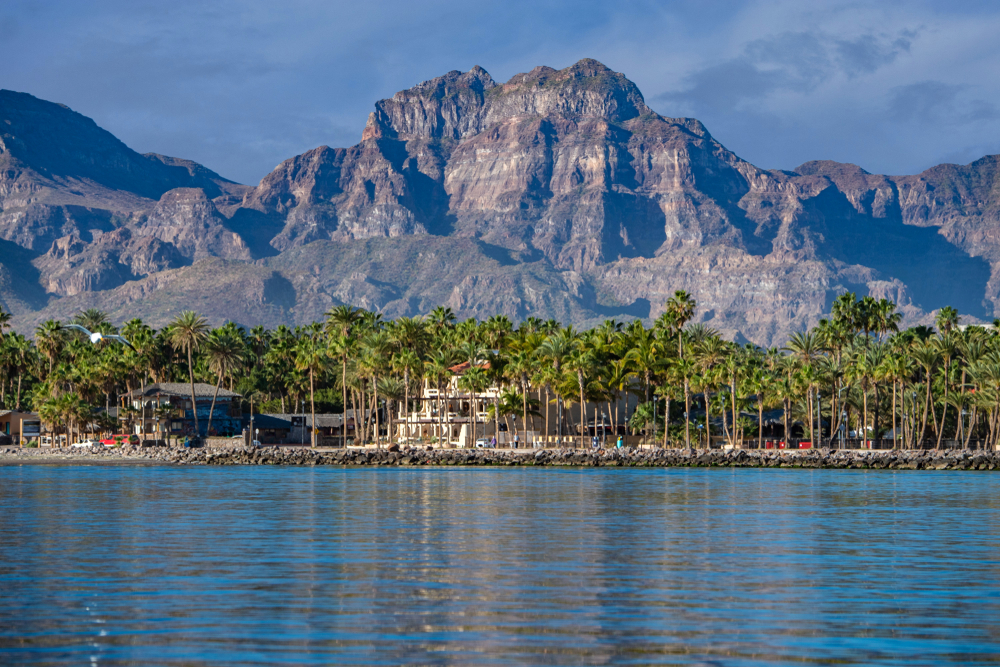 Coast of Loreto Mexico, a top pick for the best places to go in March in the world, with a towering rock face overlooking a few homes on the still water