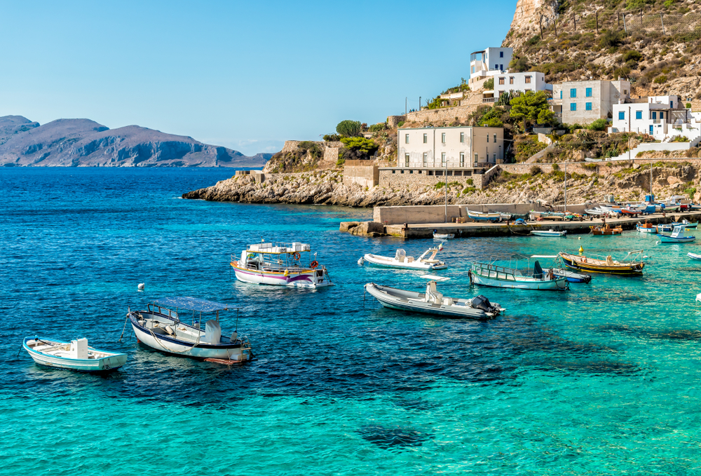 Levanzo Island pictured on a clear day with teal water in the bay and the cliffside town of Trapany, one of the best places to visit in Sicily, seen on the right