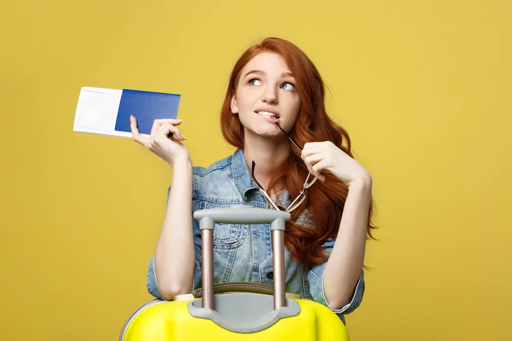 Redhead woman looks confused holding passport and flight tickets in front of her yellow suitcase for a frequently asked questions section on how to get paid to travel