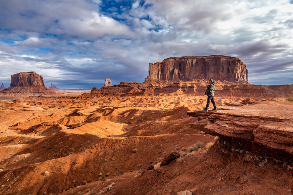 Man walking along a rock formation during the best time to visit Monument Valley with blue skies and brown dirt below him