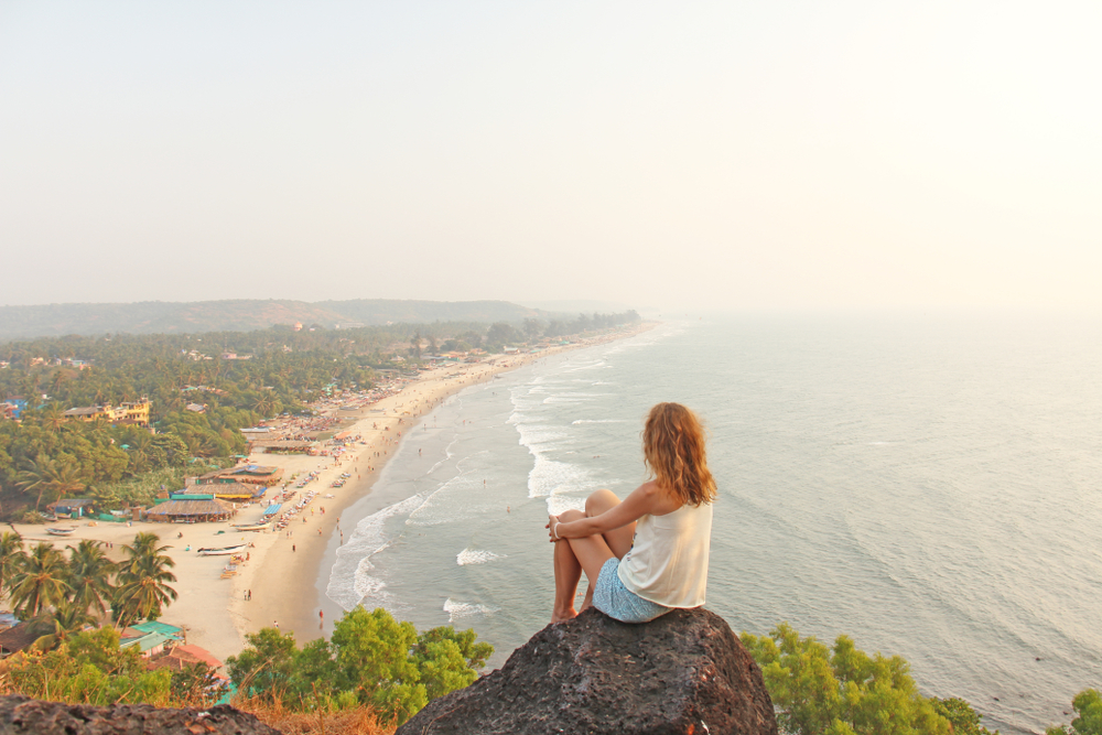 Woman sitting on a rock in Goa overlooking the beach during the best time to visit with a hazy sky above the ocean and beach