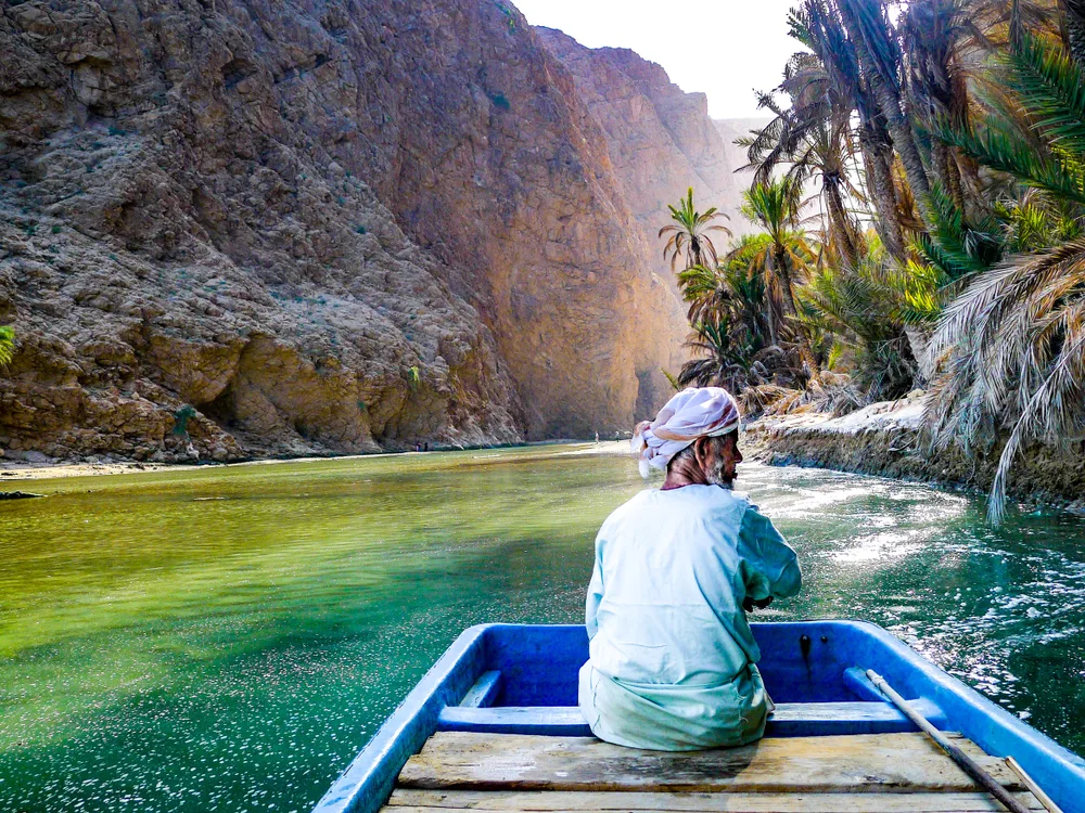 Person wearing a headscarf and a white robe sitting on a blue boat while floating along a green river during the best time to visit Oman